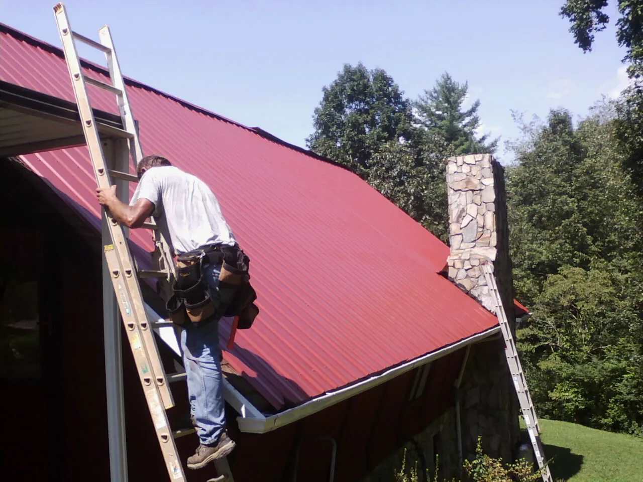 Roofer on a ladder beside a red metal roof and stone chimney — Soffit & Fascia Repair by Roofing Brothers in Touchet